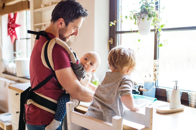 Vater mit einem Baby in einer Tragetasche und einem Kleinkind auf einem Stuhl